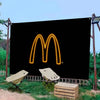 Une bannière noire avec le logo 'M' de McDonald's jaune brillant, en extérieur avec de la verdure et des palettes en bois.
