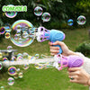 A hand is using a purple automatic bubble gun to blow bubbles into the air in a green outdoor setting.