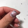 A hand holds a fingernail painted with iridescent silver and black metallic foil flakes for nail art.