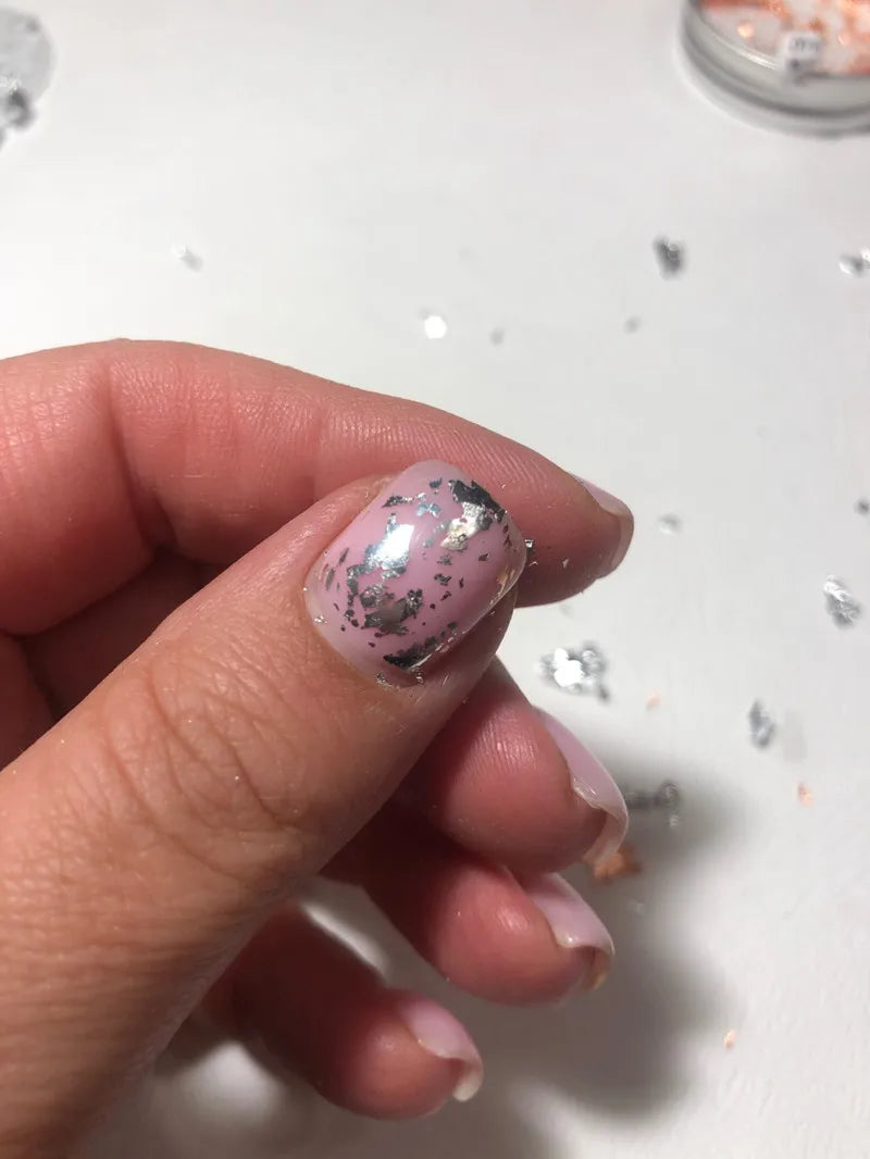 A hand holds a fingernail painted with iridescent silver and black metallic foil flakes for nail art.
