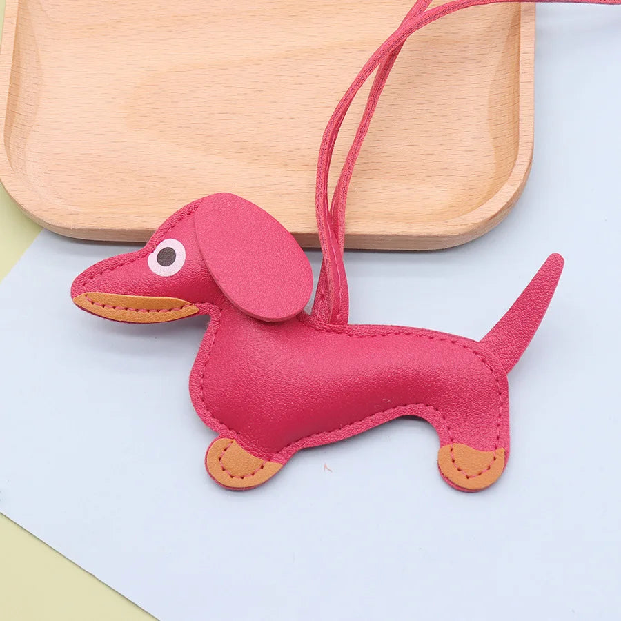 A pink dachshund puppy keychain made of PU leather lying on a white surface next to a wooden tray.