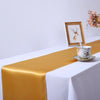 Elegant gold satin table runner draped on a white tablecloth with a small vase and framed art.