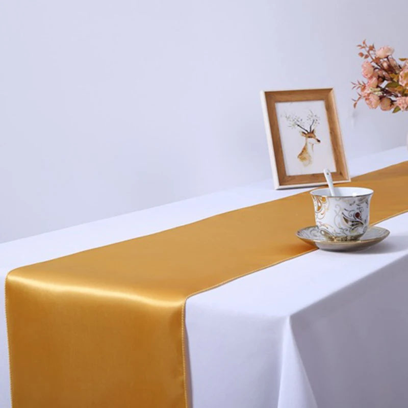 Elegant gold satin table runner draped on a white tablecloth with a small vase and framed art.