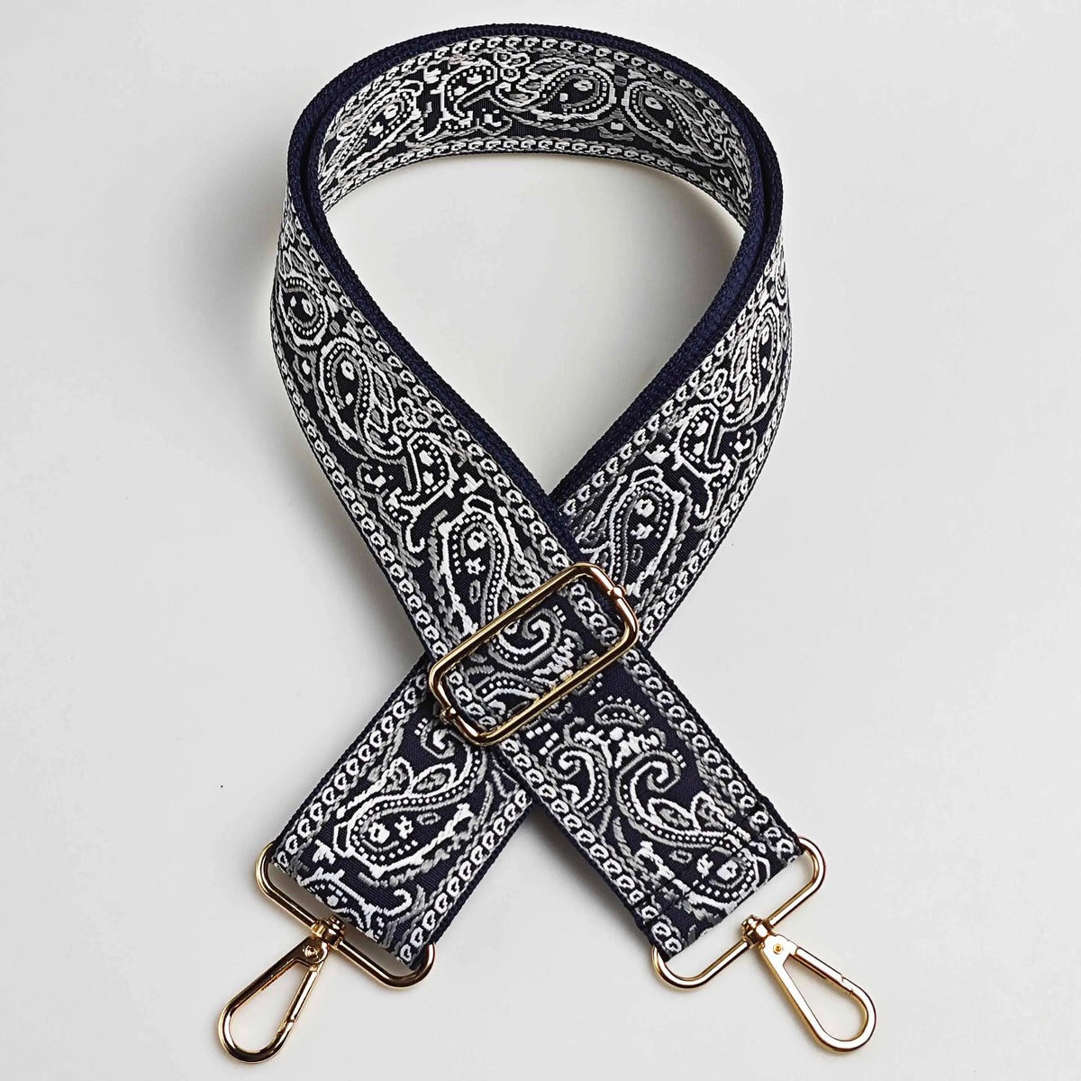 A dark navy and white paisley jacquard bag strap with a gold buckle and leather accents.