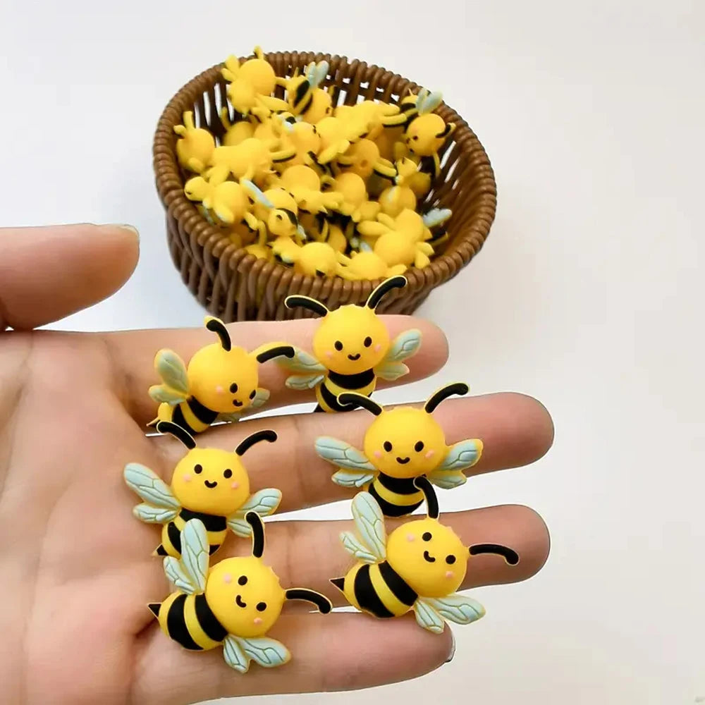 A hand holds several yellow 3D bee silicone beads and a bowl full of similar bees for jewelry making