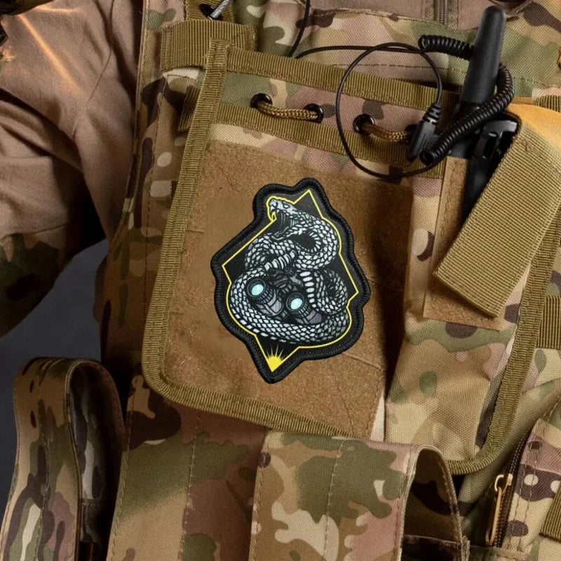 A tactical snake morale patch with a black and yellow design is attached to a camouflage fabric item.