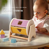 A wooden shape rolling drum toy for babies with various colored inserts and a baby looking at it.