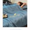 A light blue ultra-soft comforter with gold embroidery is draped over a bed, with a person resting their head on it.