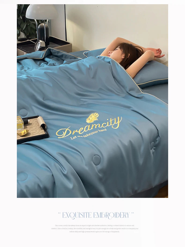 A light blue ultra-soft comforter with gold embroidery is draped over a bed, with a person resting their head on it.