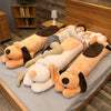 A woman is relaxing on a gray surface surrounded by several large, soft, orange and beige dog plush pillows.
