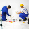 Two workers in blue coveralls and yellow hard hats are applying static-cling clear plastic sheeting to a white wall.