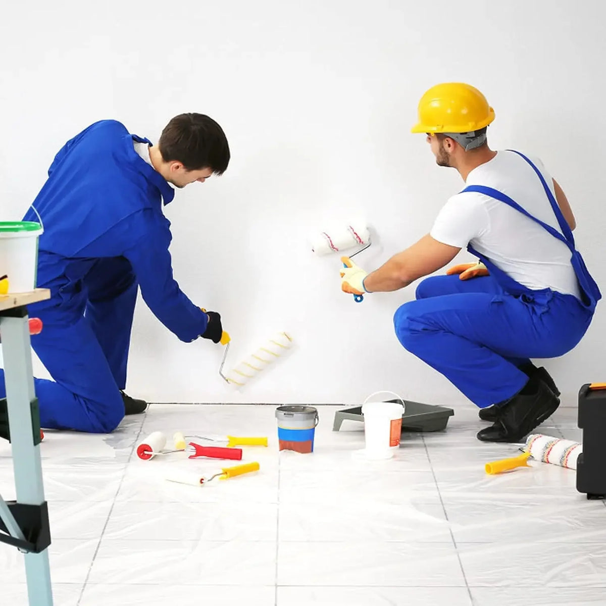 Two workers in blue coveralls and yellow hard hats are applying static-cling clear plastic sheeting to a white wall.