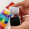 A hand holds a spool of white stretchy elastic cord next to various colorful elastic cords for jewelry making.