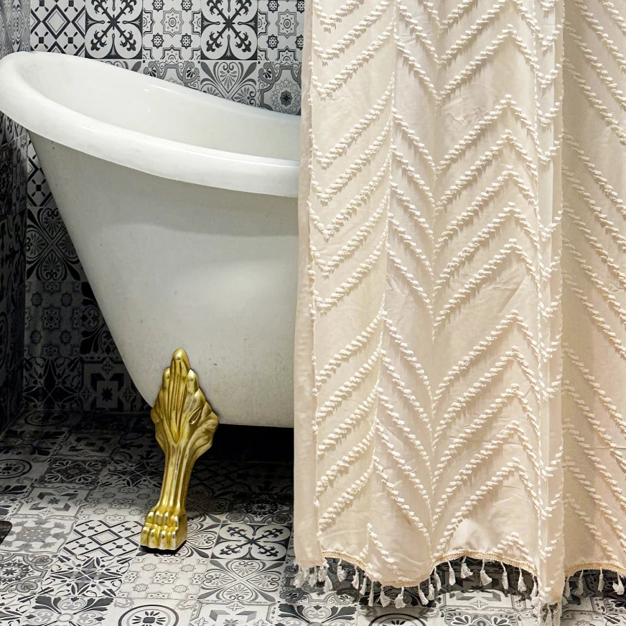 A beige bohemian floral cutout tassel shower curtain hangs next to a white bathtub and gold fixture on patterned tile floor.