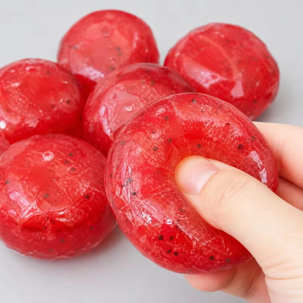 Une main tient un jouet anti-stress en forme de pastèque rouge avec des graines noires visibles.