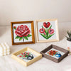 Three framed cross stitch designs featuring roses and tulips on white fabric displayed on a white surface.