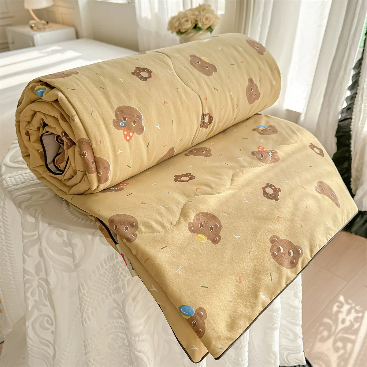 A tan quilt with a pastoral print featuring small brown teddy bears and yellow accents, rolled up on a white bed.