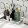 A set of glass soap dispensers with gold pumps and a small black bottle displayed on a white marble tray against a hexagonal 