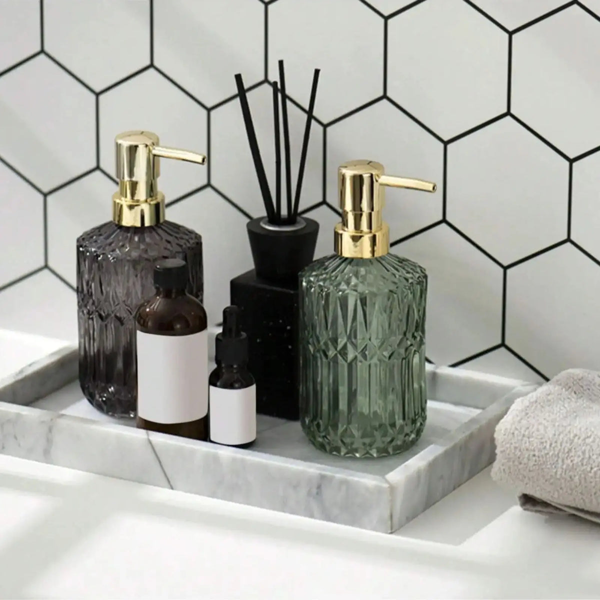 A set of glass soap dispensers with gold pumps and a small black bottle displayed on a white marble tray against a hexagonal 