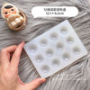 A silicone paint palette tray with twelve wells is shown next to a small doll and a hand on a gray furry surface.