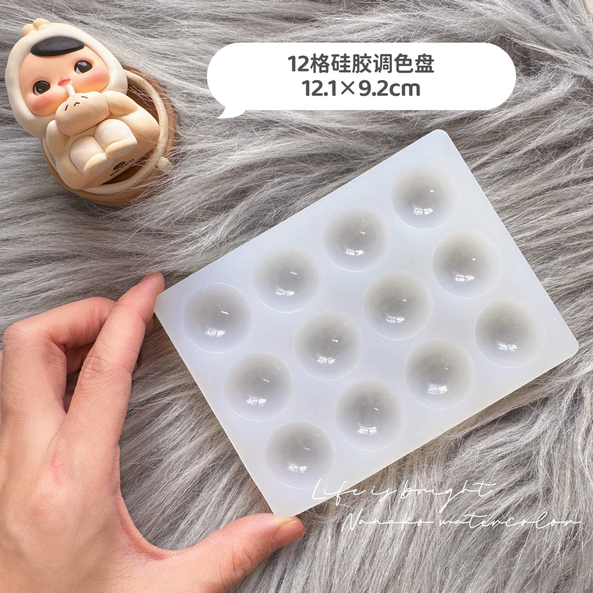 A silicone paint palette tray with twelve wells is shown next to a small doll and a hand on a gray furry surface.