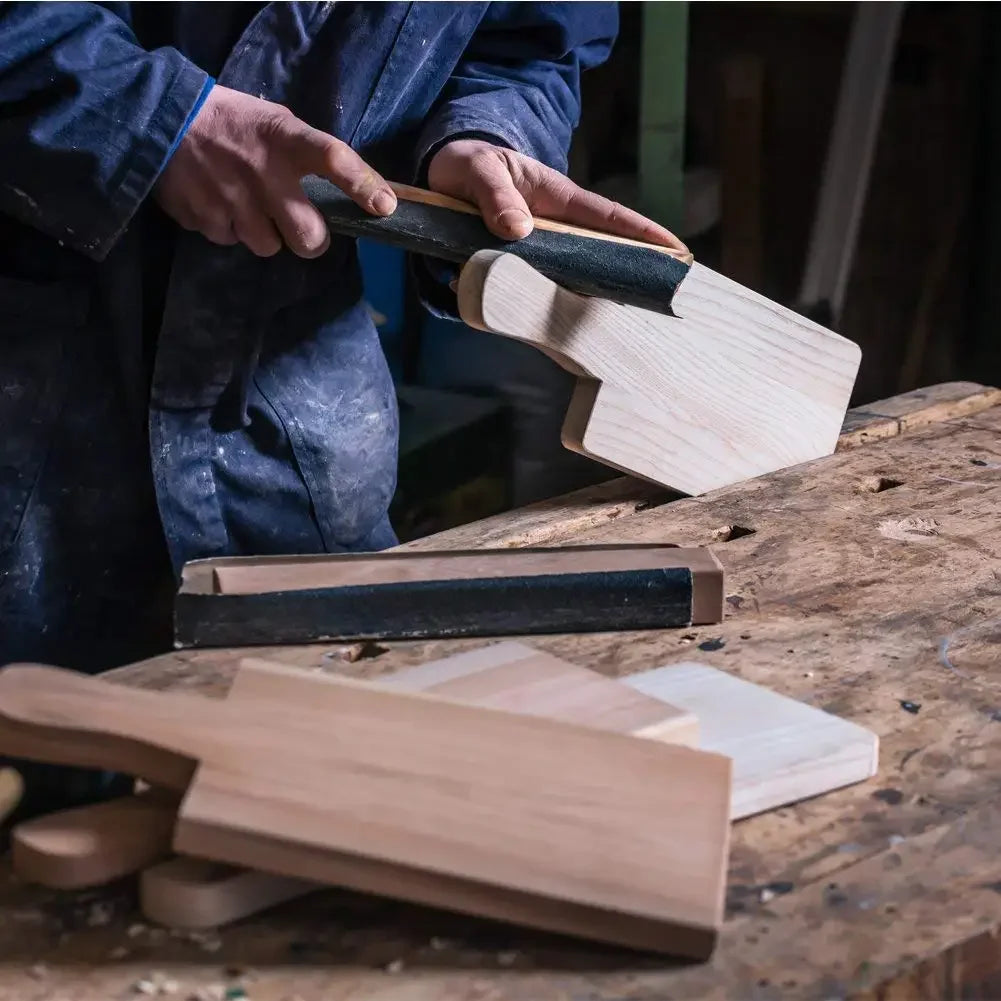 A person is using a dark-handled sanding block to sand a piece of light wood on a wooden surface.