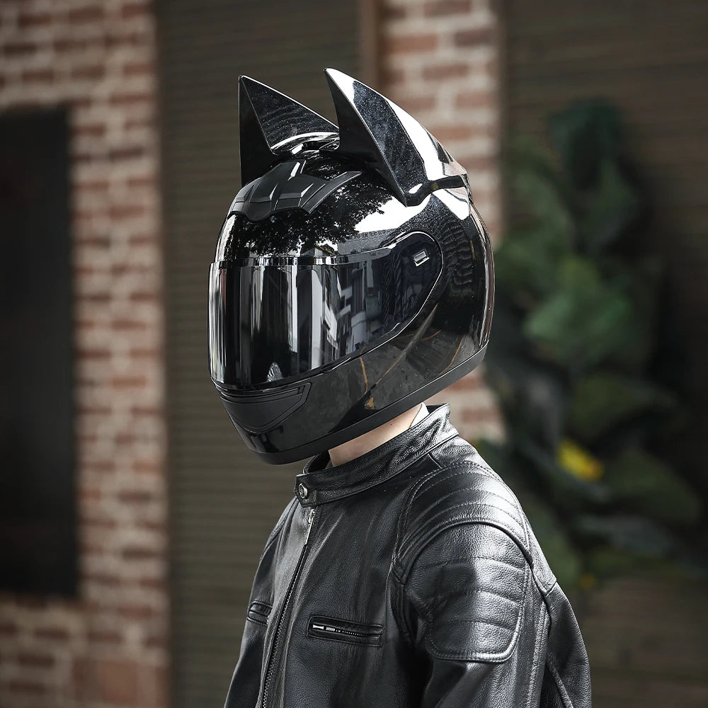 A person wearing a black bat-themed full-face motorcycle helmet with detachable horns, standing in front of a brick wall.