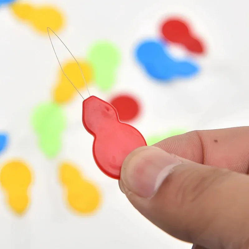 A hand holds a red plastic needle threader with a pointed tip against a background of colorful, irregularly shaped plastic pi