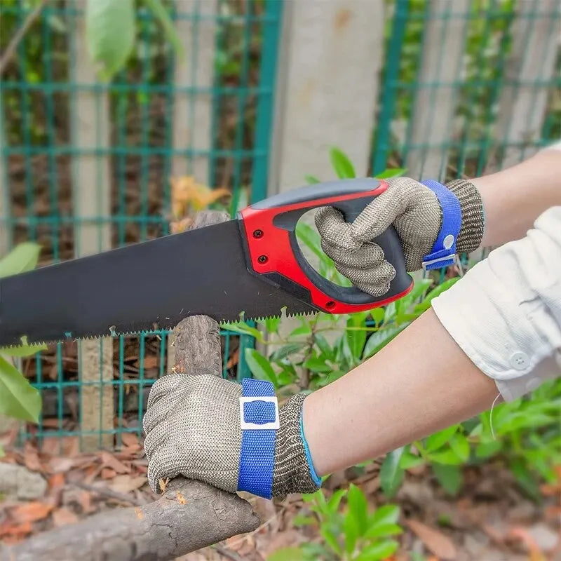A hand wearing safety gloves is using a black knife to cut a piece of wood outdoors.