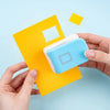A hand holds a yellow embossing punch and a light blue stamp on a light-colored card against a light blue background.