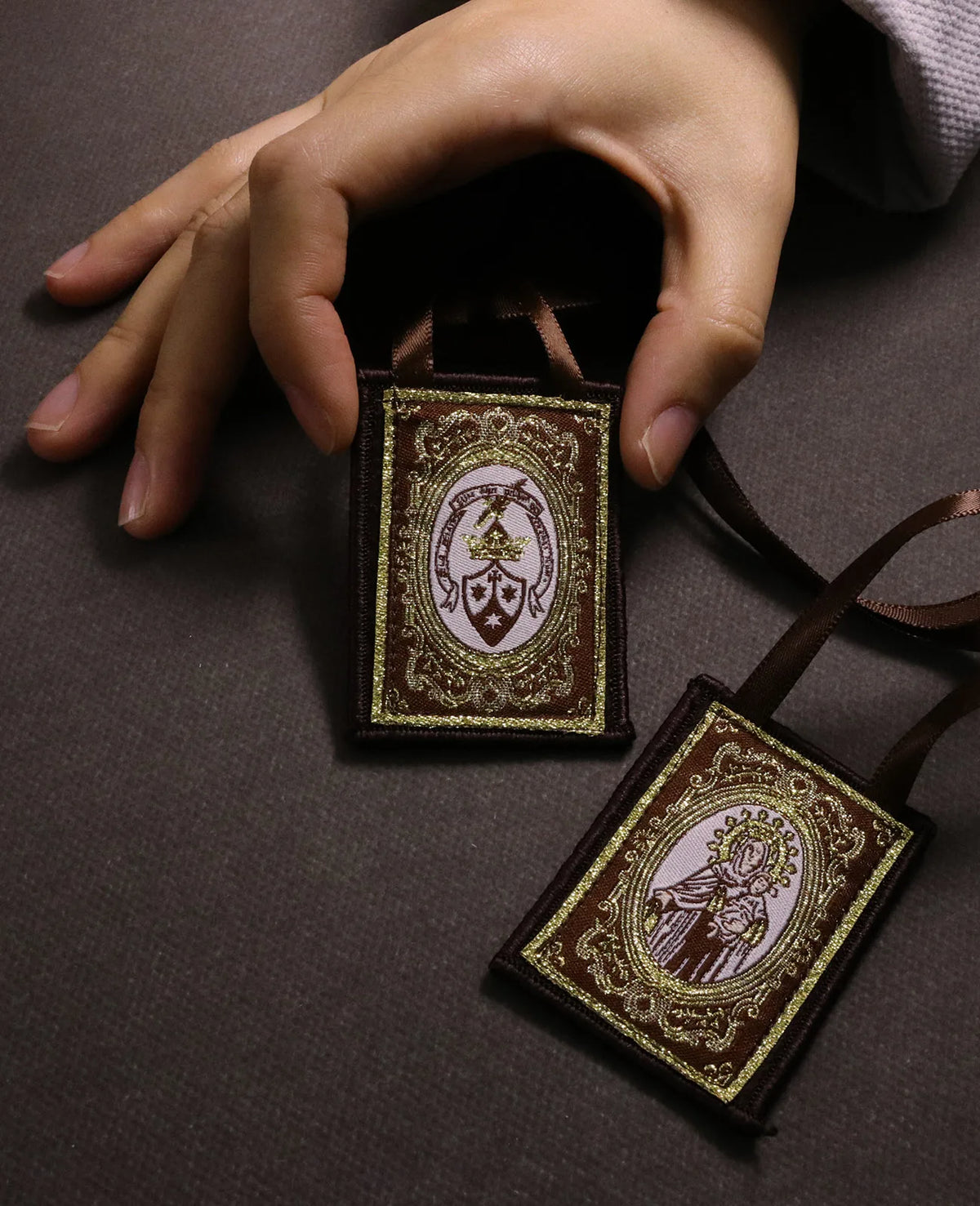 A hand holds a brown fabric scapular necklace with an ornate pendant against a dark background.