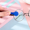 A hand holds a blue cleaning brush attached to a pair of eyeglasses on a pink surface.
