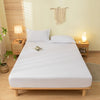 A white fitted bed sheet set with pillowcases is displayed on a wooden bed frame in a bright bedroom.