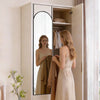 A woman stands in front of a full length wall mirror with an arched frame, looking at her reflection.