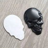 A black 3D chrome skull emblem badge is shown next to its white, unprinted counterpart on a gray surface.