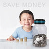 A smiling boy sits next to a clear glass jar filled with silver coins on a white surface.
