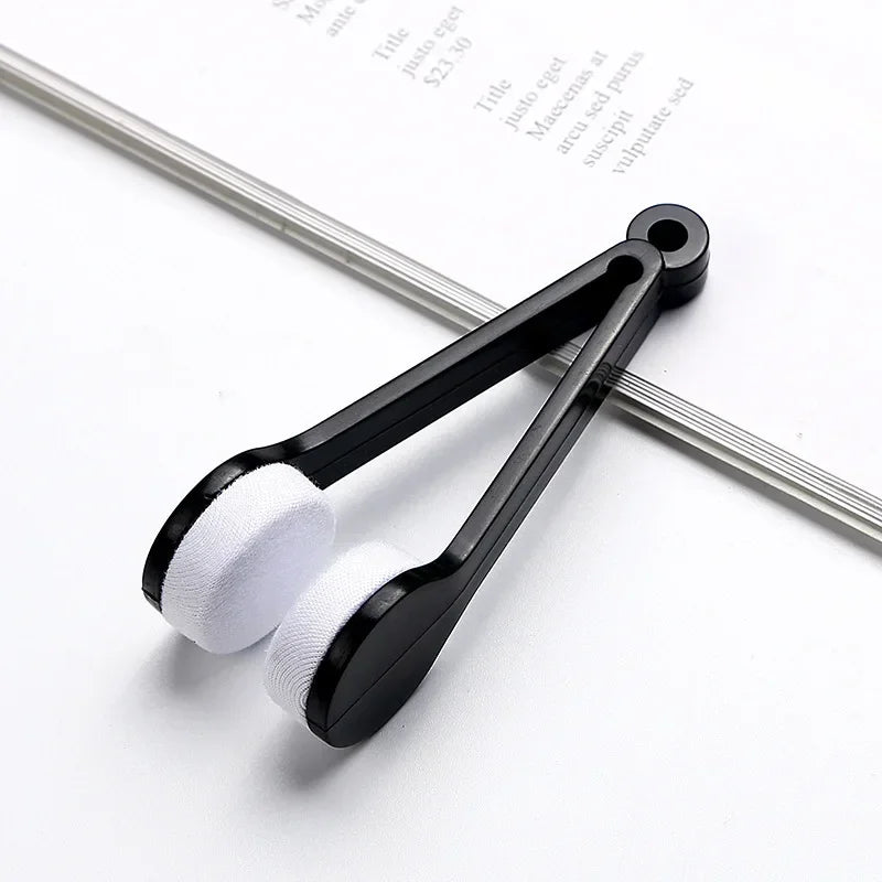 A black portable double-sided glasses cleaning brush with white microfiber pads resting on a sheet of paper.