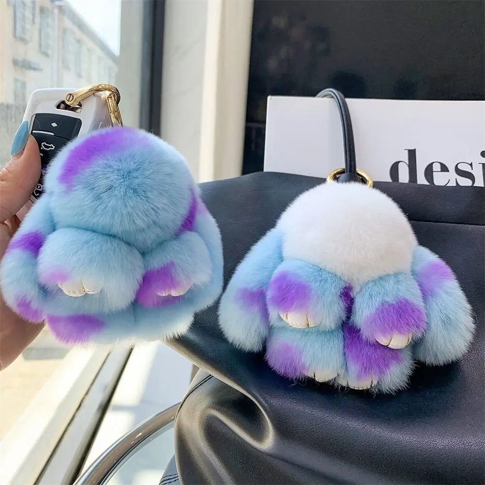 A soft plush bunny keychain featuring blue and purple fur with an ivory accent, held by a hand.