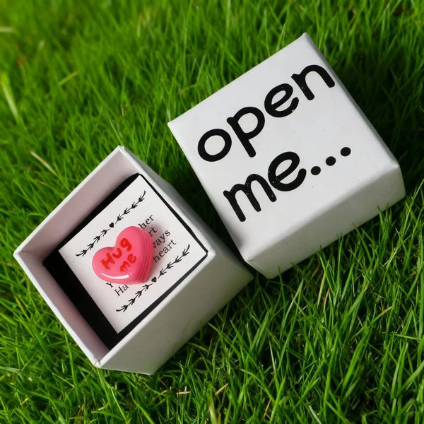 A white gift box with the text open me displayed on green grass, featuring a pink resin heart and love card.