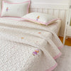 A white quilted bedspread with pink trim and small purple floral embroidery is displayed on a bed with matching pillows.
