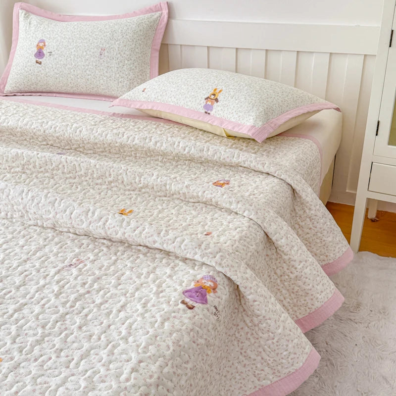A white quilted bedspread with pink trim and small purple floral embroidery is displayed on a bed with matching pillows.