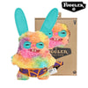 A fluffy, multi-colored plush toy monster with large blue eyes and funny teeth is shown next to its packaging box.