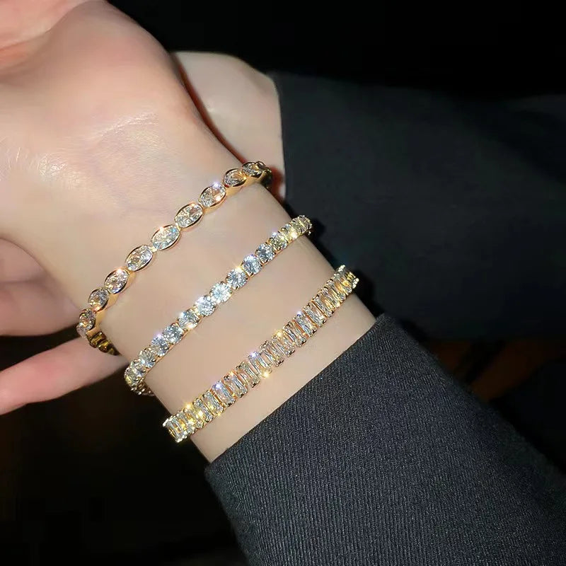 Nahaufnahme einer Hand mit drei gestapelten, zarten goldenen Tennisarmreifen mit Diamant-Steinen.