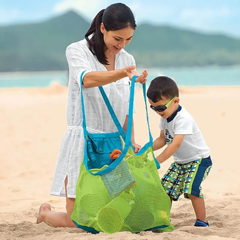 A woman is holding a large mesh green beach tote bag while interacting with a young boy on a sandy beach.