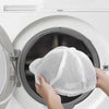 A hand places a white mesh laundry bag for hats and delicate clothing into the drum of a white washing machine.