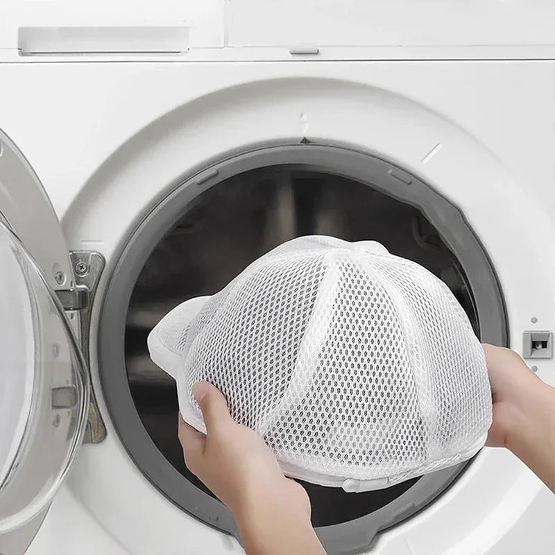 A hand places a white mesh laundry bag for hats and delicate clothing into the drum of a white washing machine.