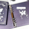 A close-up of a silver butterfly design on a purple photo album page with an ornate metal butterfly clip.
