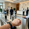 A hand is interacting with a silver access control reader device in a modern building lobby.