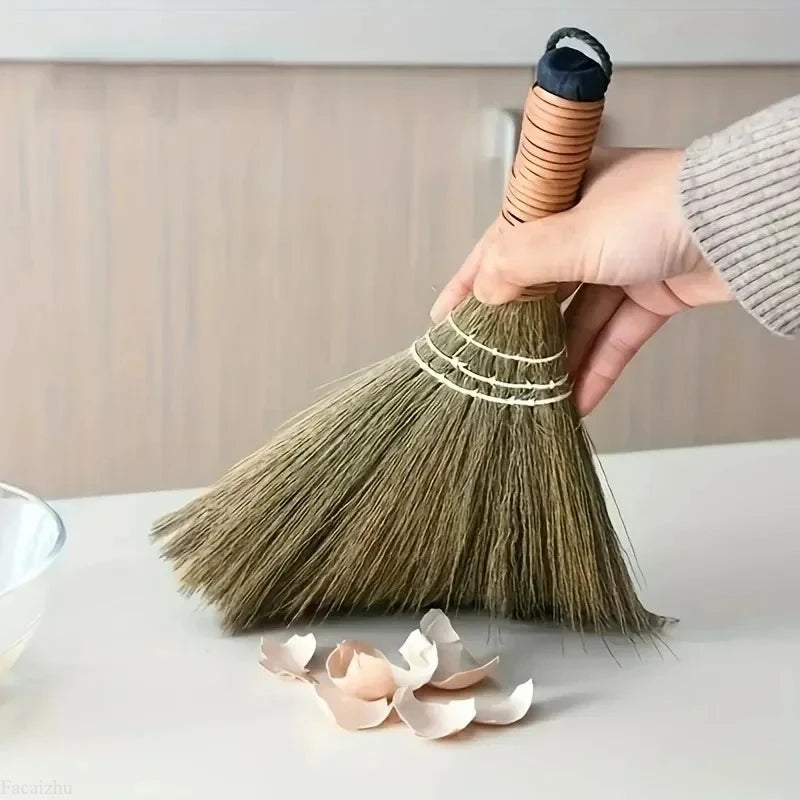 A hand is using a natural grass broom to sweep a surface, showing the bristles and handle.