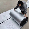 A person is rolling a roll of white automotive sound deadening and heat insulation mat on a gray floor.